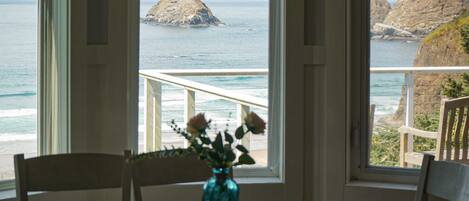 A room with a large window offering a view of the ocean and rocky cliffs. There is a wooden table with a vase of flowers in the center.