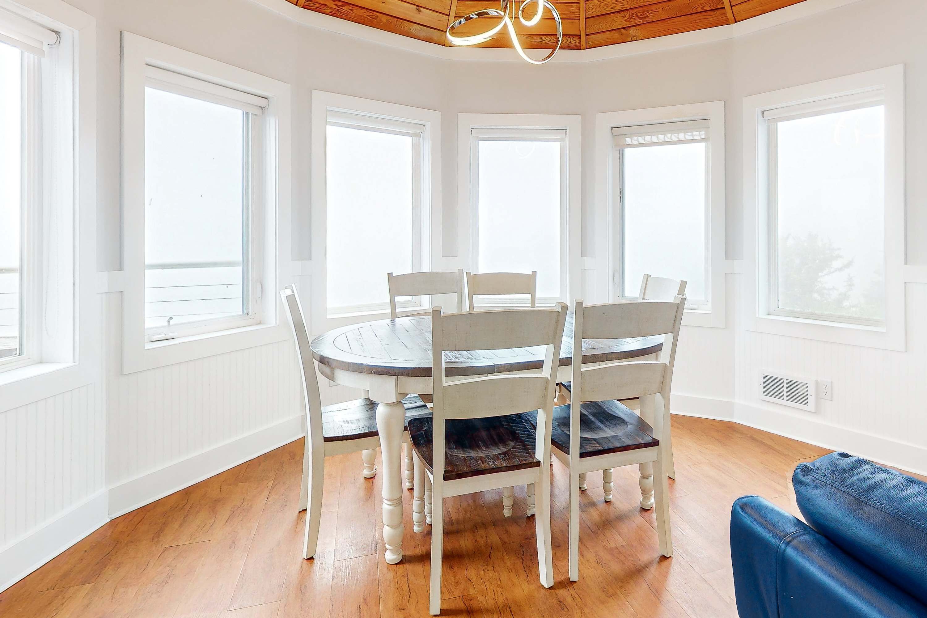 Un área de comedor con una mesa redonda y sillas blancas, ventanas amplias y un sofá azul.