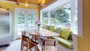 Un coin repas avec une table et des chaises en bois, un canapé avec des coussins verts et une vue sur les arbres à travers de grandes fenêtres.