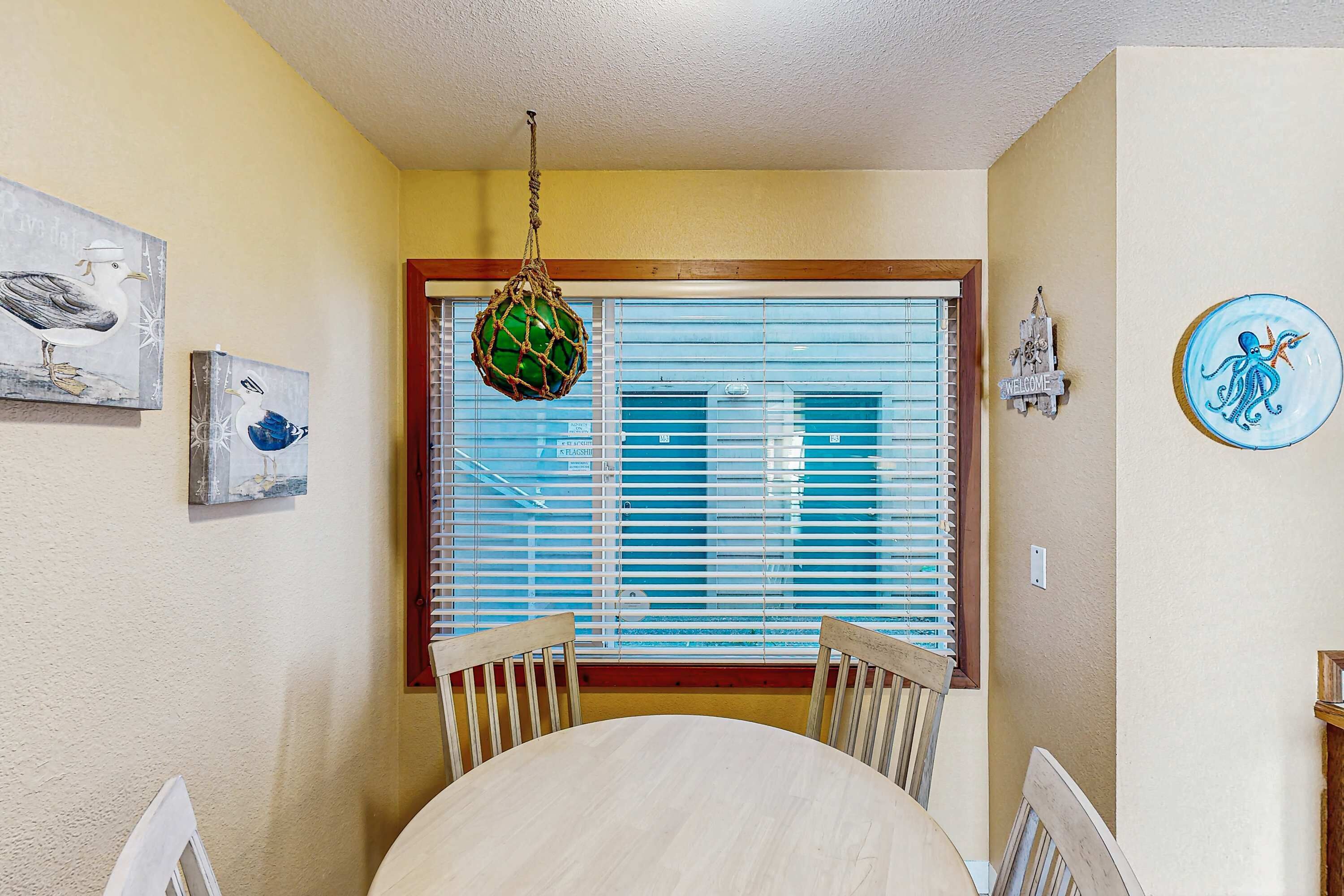 A dining area with a round table, two wooden chairs, a hanging green ornament, and a window with blinds.