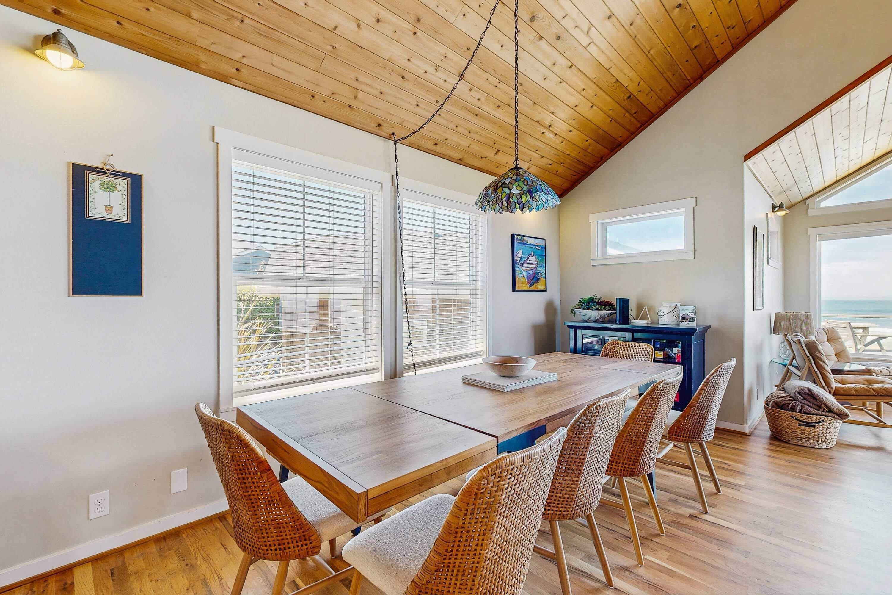 A dining area with a wooden table and wicker chairs, large windows with blinds, and a hanging light fixture.
