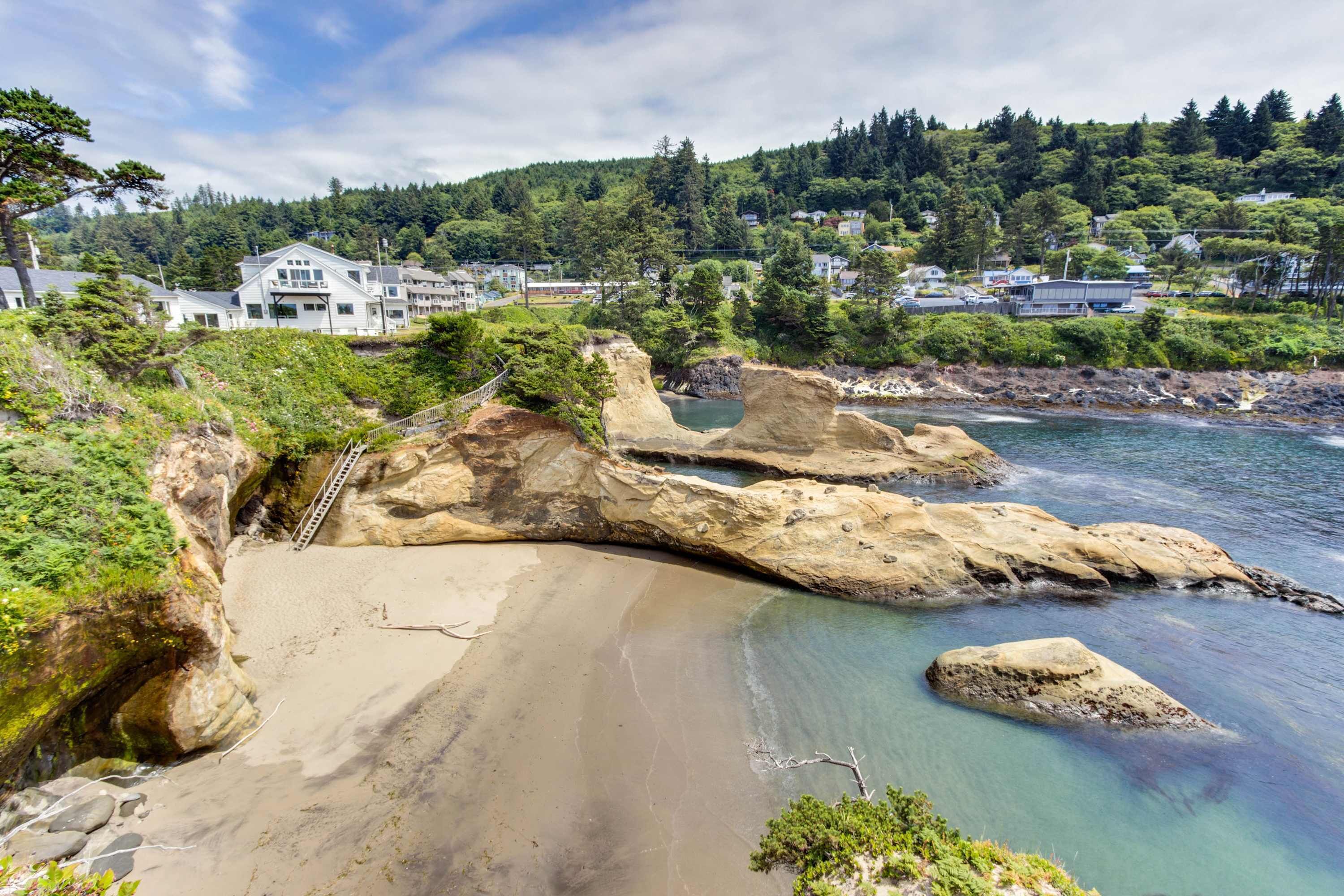 A coastal landscape with a sandy beach, rocky formations, and a residential area in the background.