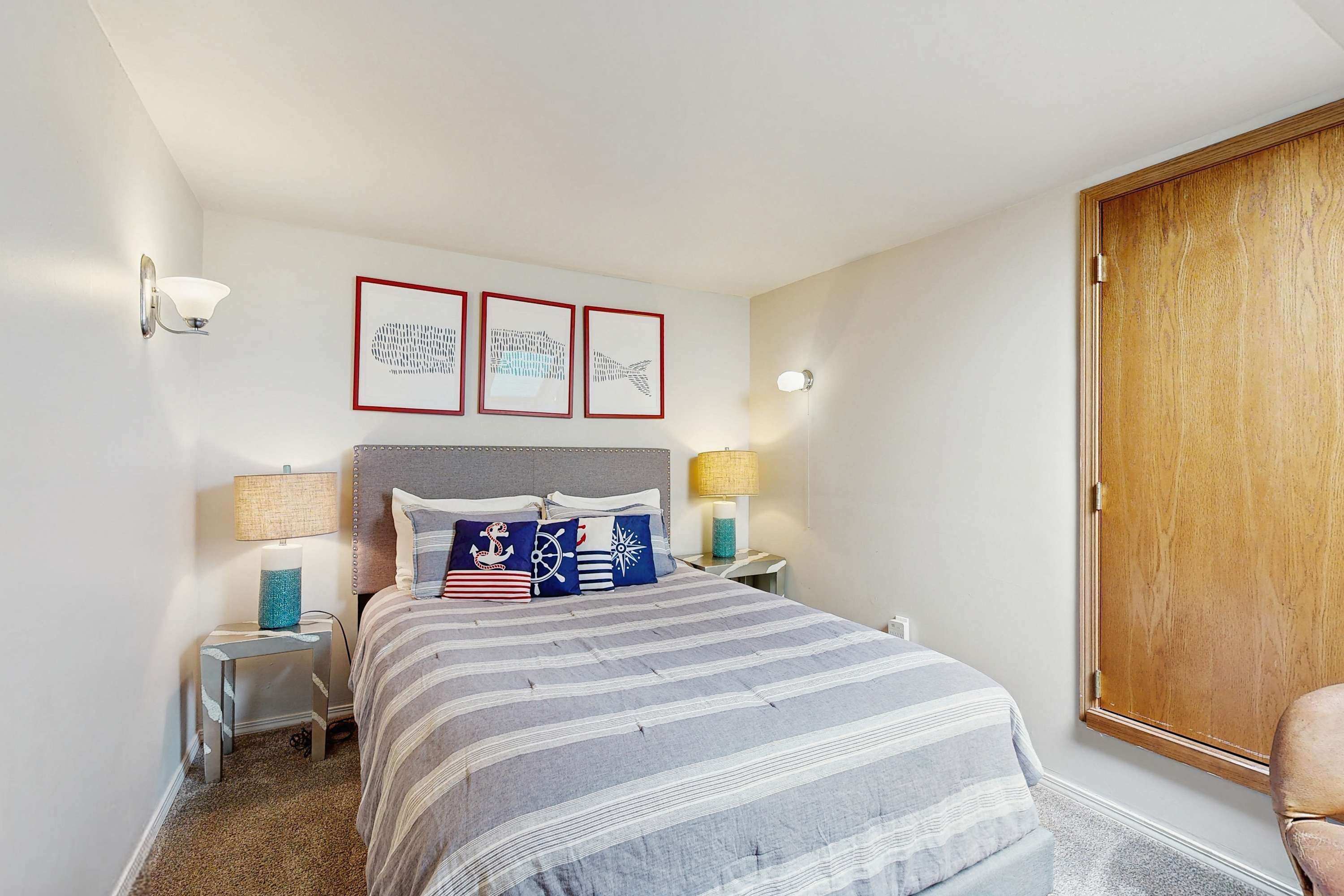 A neatly arranged bedroom with a bed, two bedside tables with lamps, a wooden door, and three framed pictures on the wall.