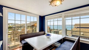 Un comedor con una mesa y bancos, ventanas amplias con vistas al mar y edificios, y una lámpara en el techo.