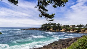 A coastal landscape with rocky terrain, ocean waves, and a line of trees and buildings in the background. - Ocean view cabin with deck and beach access dog friendly (Depoe Bay)