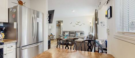 A modern kitchen with a stainless steel refrigerator, wooden dining table, and a living area with a sofa and decorative wall art.
