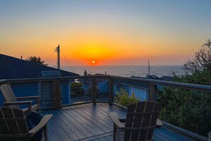 Um deck de madeira com cadeiras, com vista para o pôr do sol sobre o oceano.