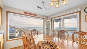 Un comedor con vistas a la playa, sillas de madera, una mesa grande y un cartel sobre las ventanas.