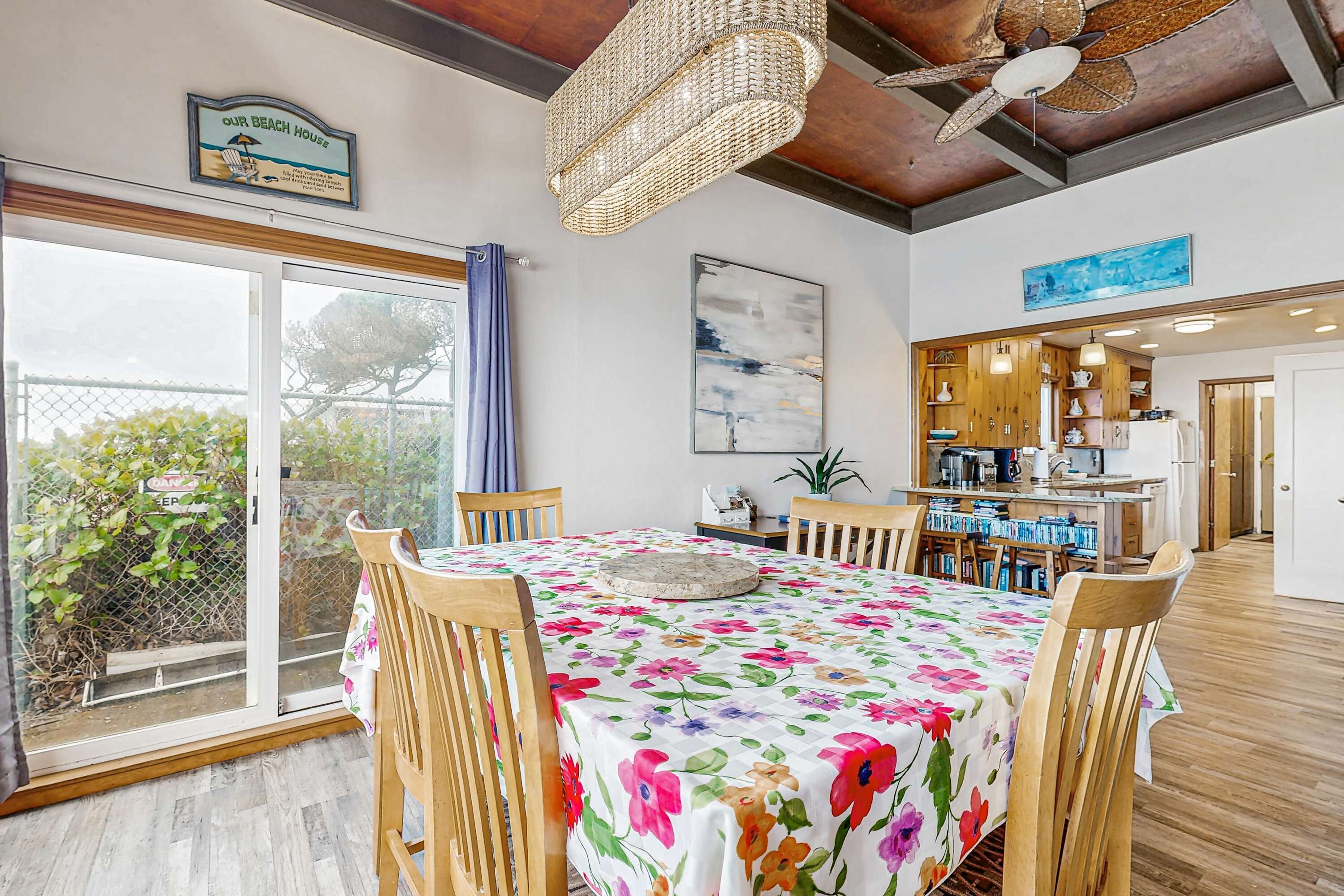 A dining area with a table covered in a floral tablecloth, wooden chairs, a chandelier, and a view of the outdoors through a glass door.