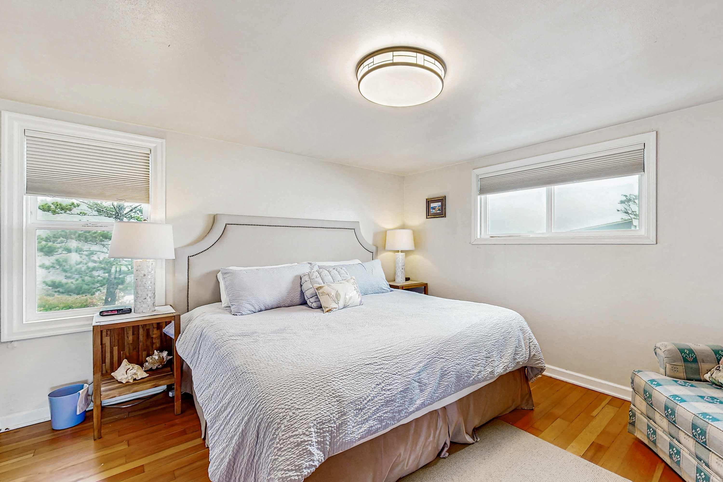 A bedroom with a large bed, two bedside tables, a nightstand, a blue trash bin, and a window with blinds.