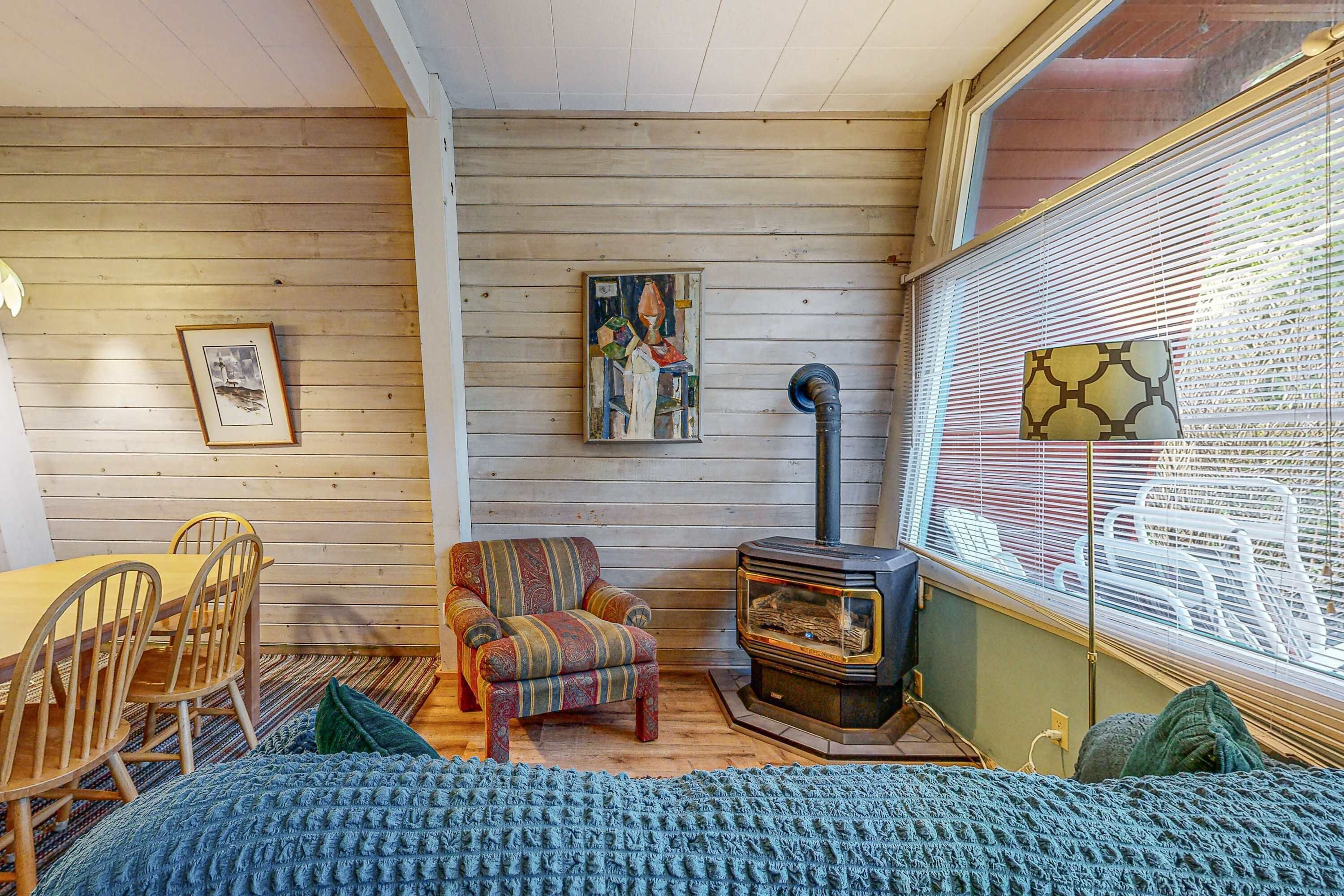 Una habitación acogedora con suelo de madera, un sillón estampado, una mesa de comedor con sillas, un cuadro colgado en la pared y un estufa de leña.