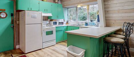 Een keuken met groene kastjes, een witte koelkast, een magnetron en een spoelbak. Er is een raam met uitzicht op bomen en een afvalbak.