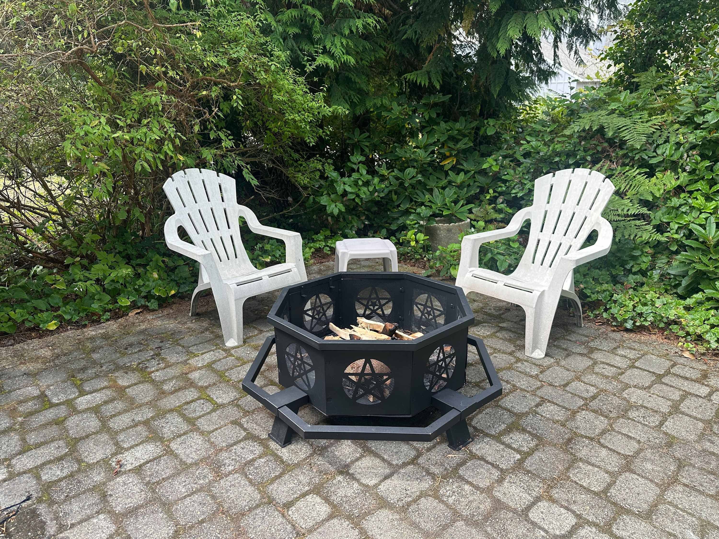 A hexagonal fire pit with logs, surrounded by two white Adirondack chairs and a small table.