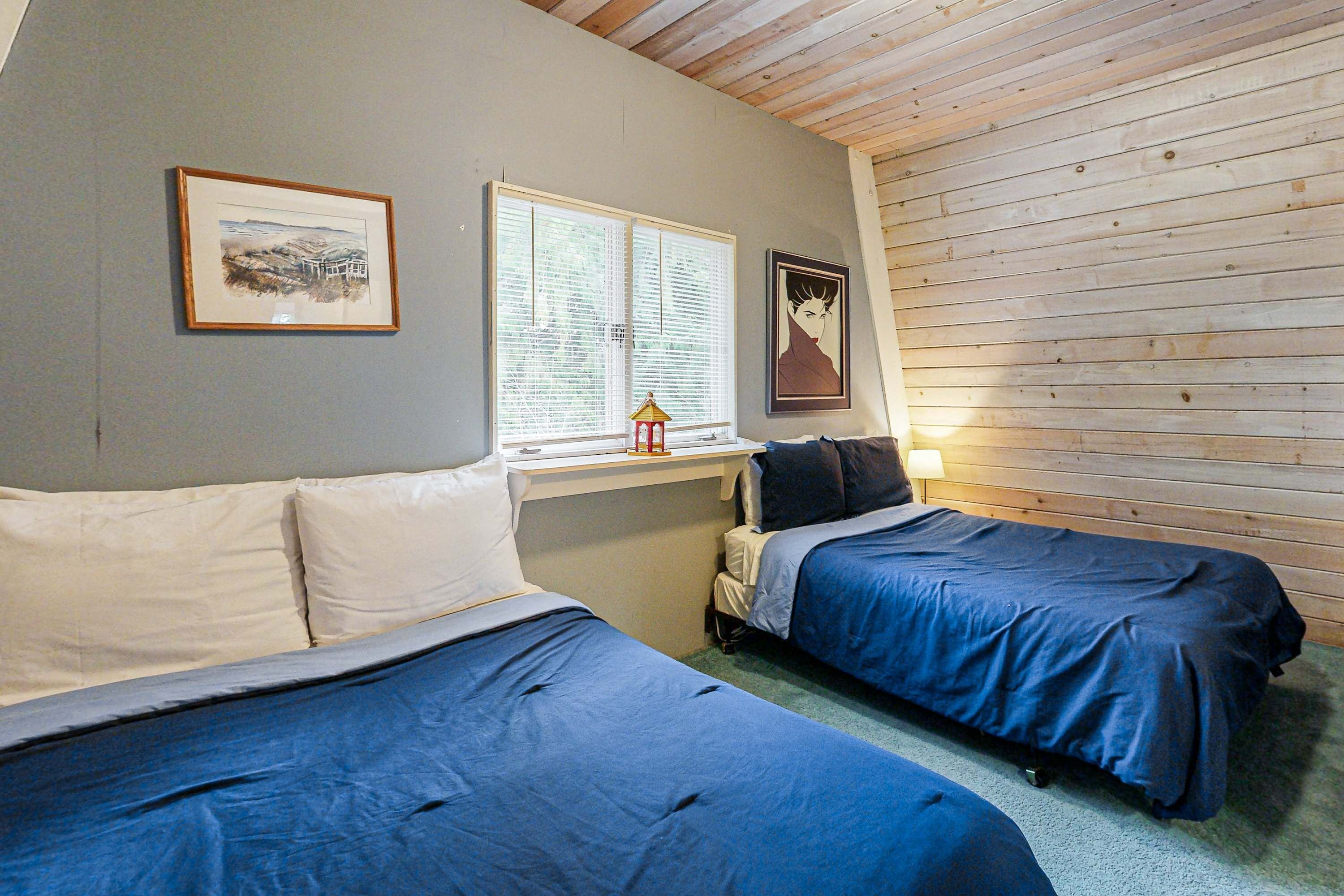 A room with two beds, a window with blinds, a small table with a lamp, and framed pictures on the wall.