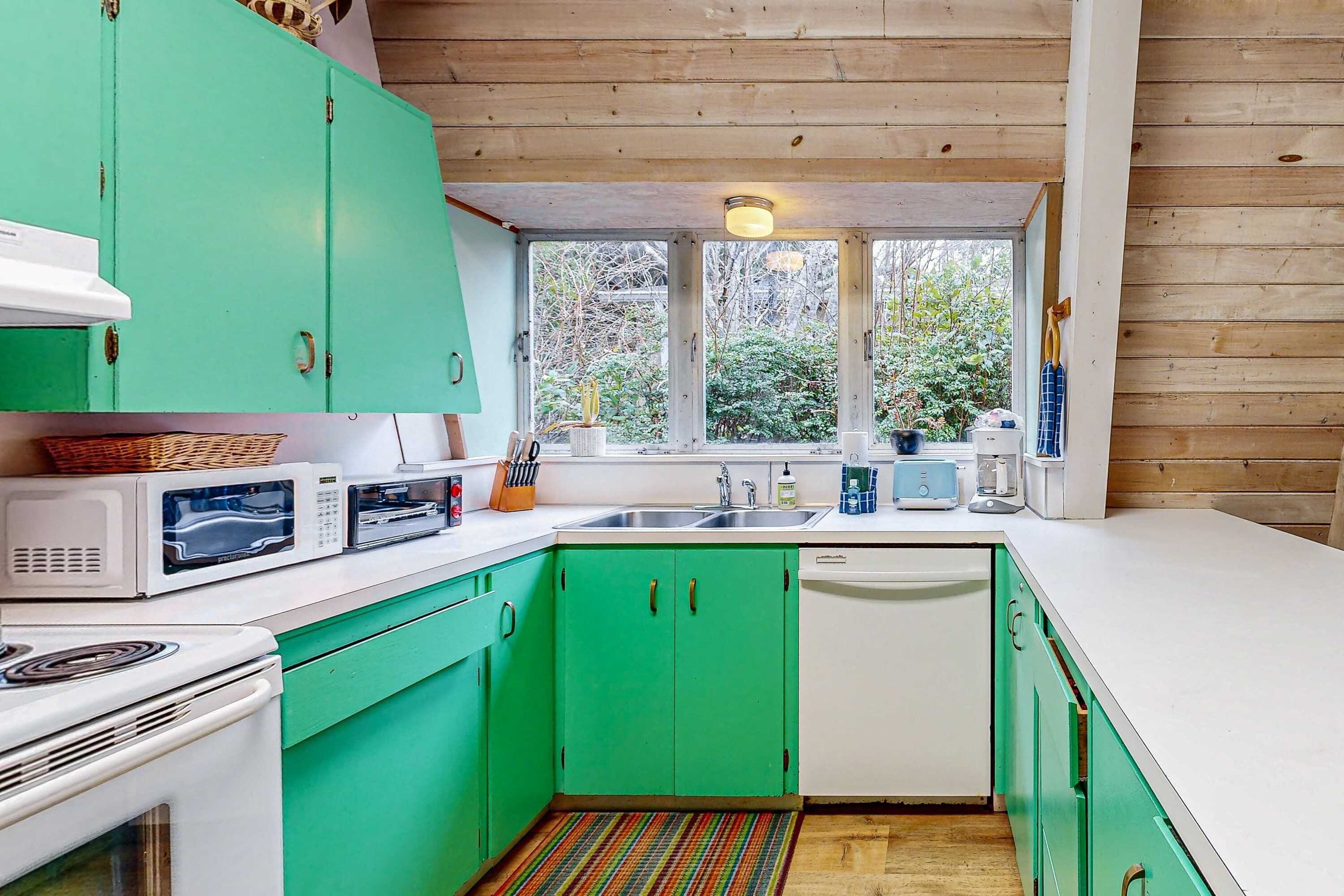 Cocina compacta con gabinetes verdes, encimeras blancas y una ventana que proporciona luz natural.