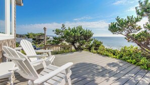 Una terraza de madera con sillas Adirondack blancas y una mesa de vidrio, con vista al mar, árboles y cielo despejado.