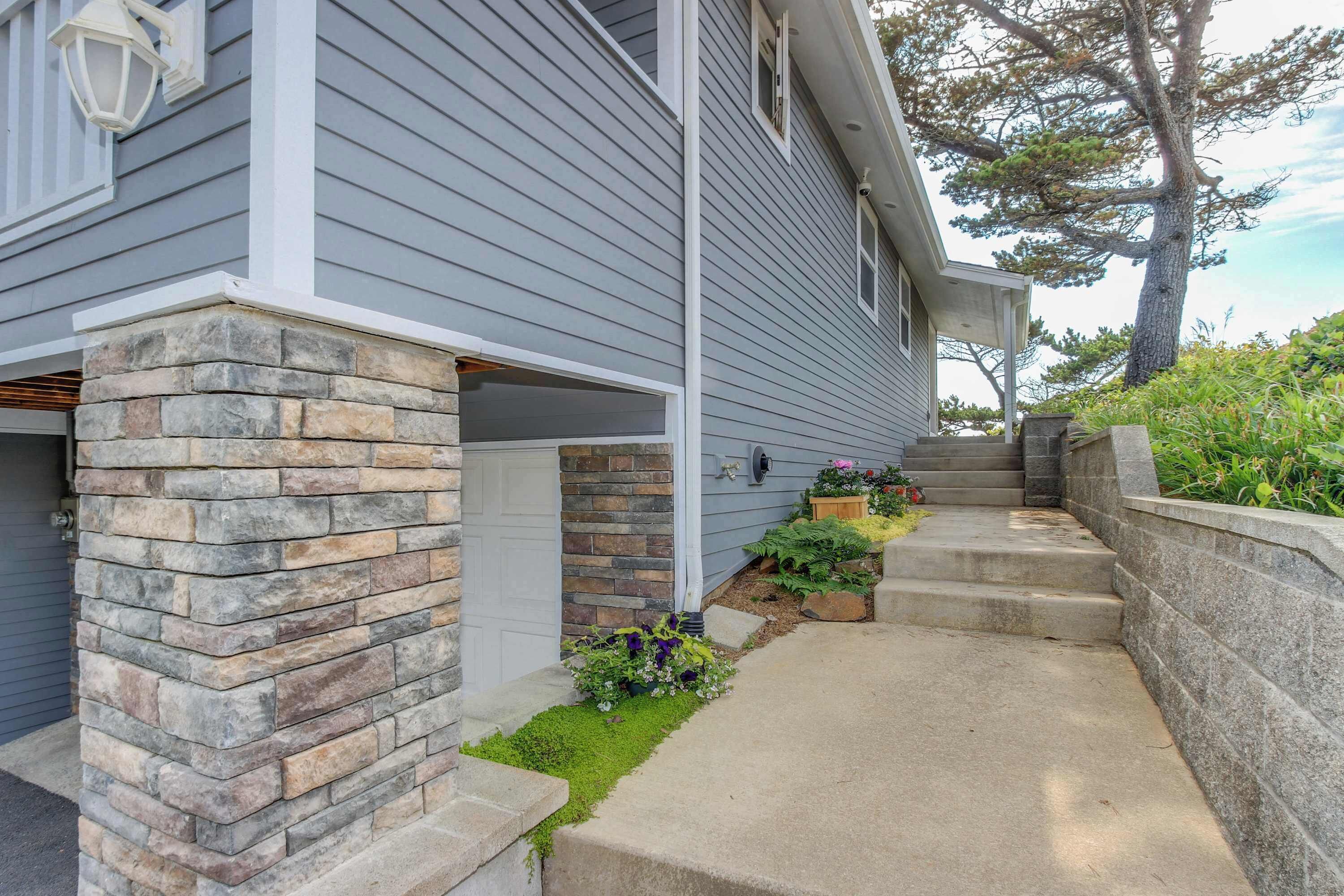 A house with a stone foundation, a concrete pathway, and a small garden with flowers.