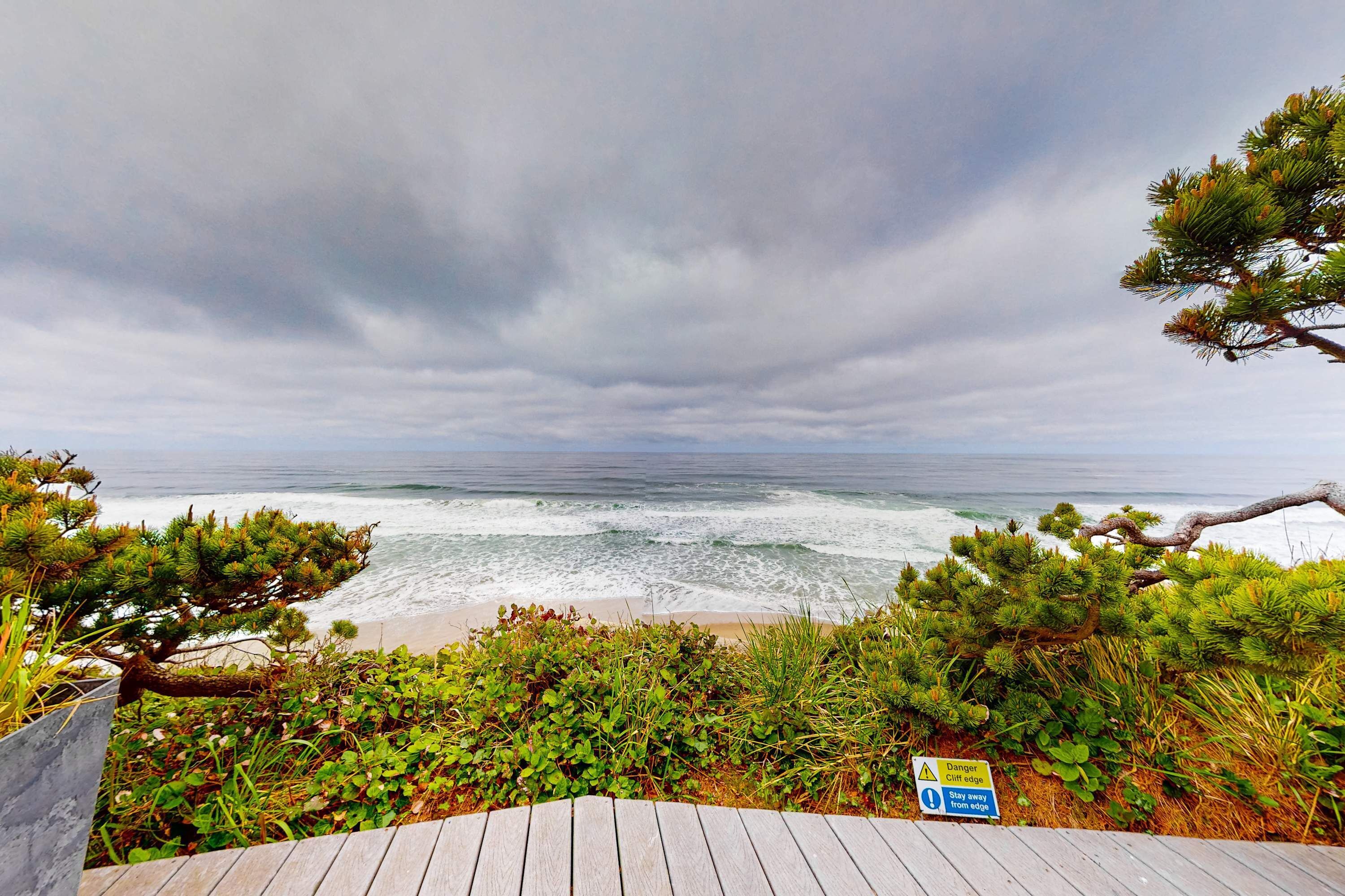 Um deck de madeira com vista para uma praia, com placa de aviso sobre beira de penhasco.