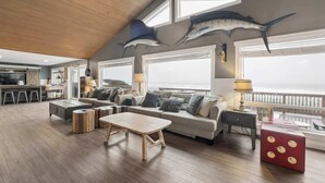 Een ruime woonkamer met een hoge plafond, houten vloer, een zithoek, een salontafel en grote ramen met uitzicht op zee.