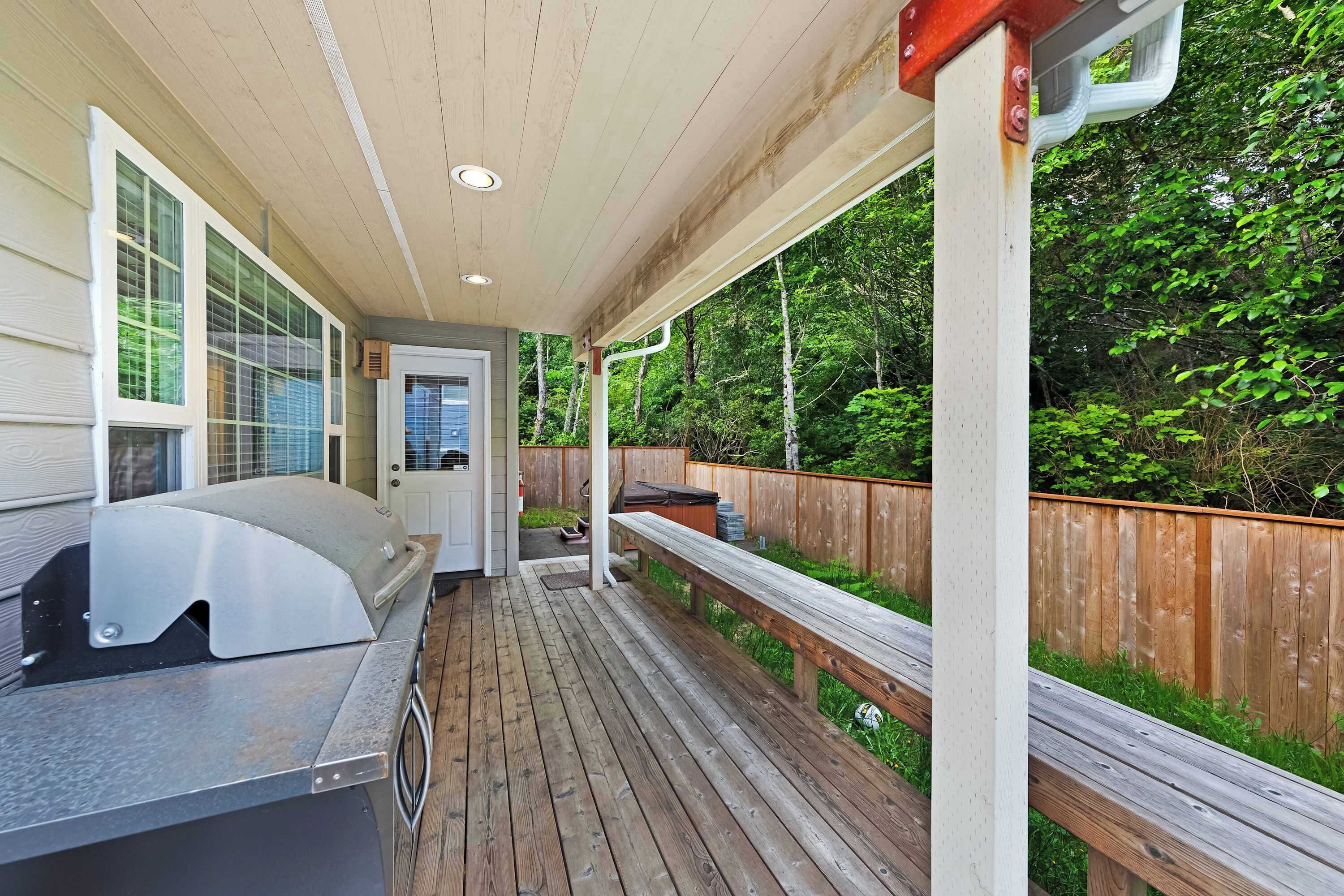 A covered outdoor deck with a grill, wooden railings, and a view of a wooded area.