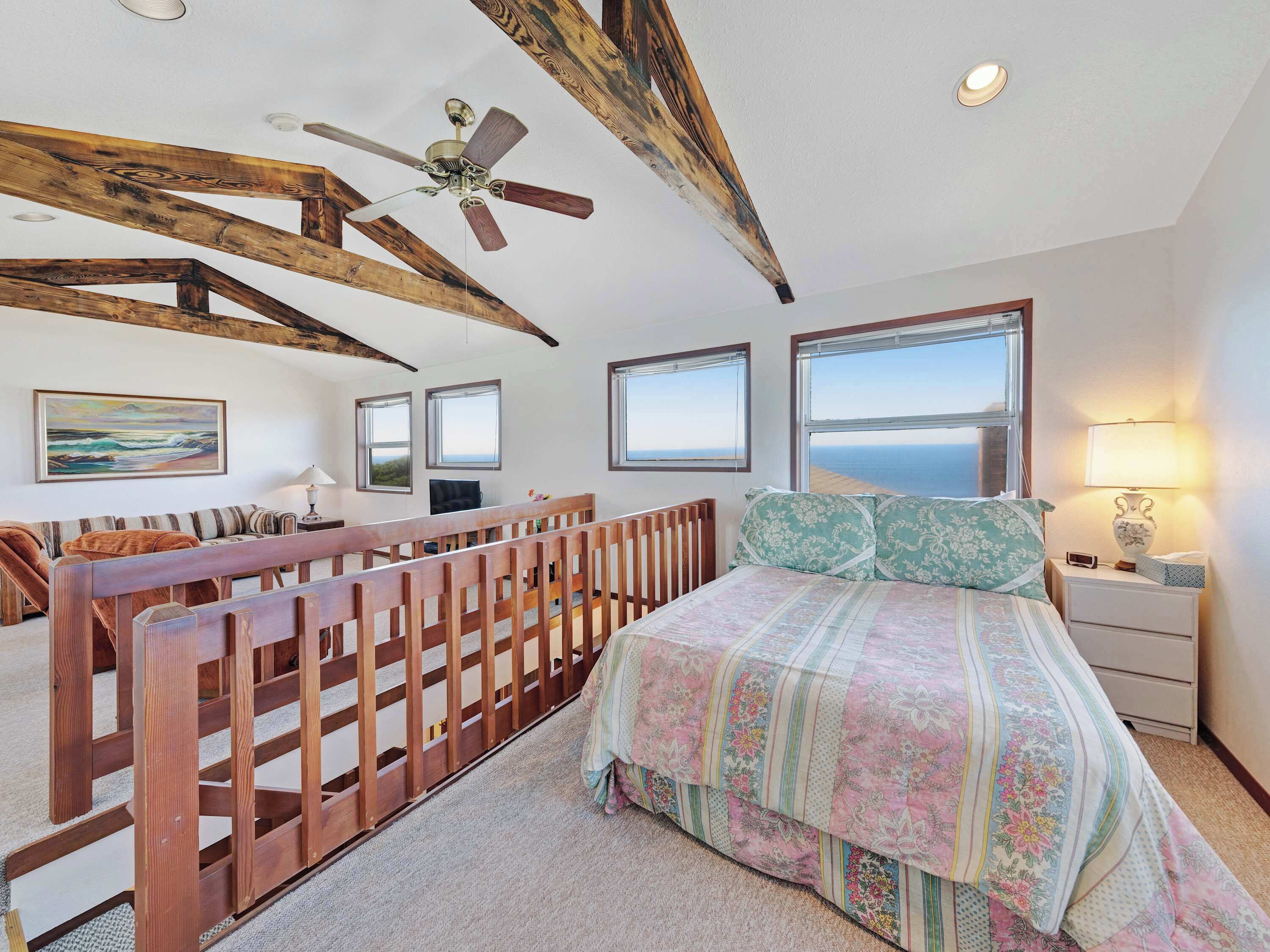 A bedroom with a wooden ceiling, a bed with floral bedding, a nightstand with a lamp, and a view of the sea through large windows.