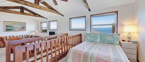 A bedroom with a wooden ceiling, a bed with floral bedding, a nightstand with a lamp, and a view of the sea through large windows.