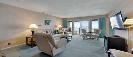 A hotel room with a living area featuring a sofa, armchairs, a coffee table, a TV, and a balcony with a view.