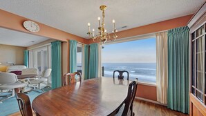 A dining room with a large wooden table, chairs, a chandelier, and a view of the ocean through large windows. - Lovely oceanfront condo with sweeping water views and balcony (Gleneden Beach)