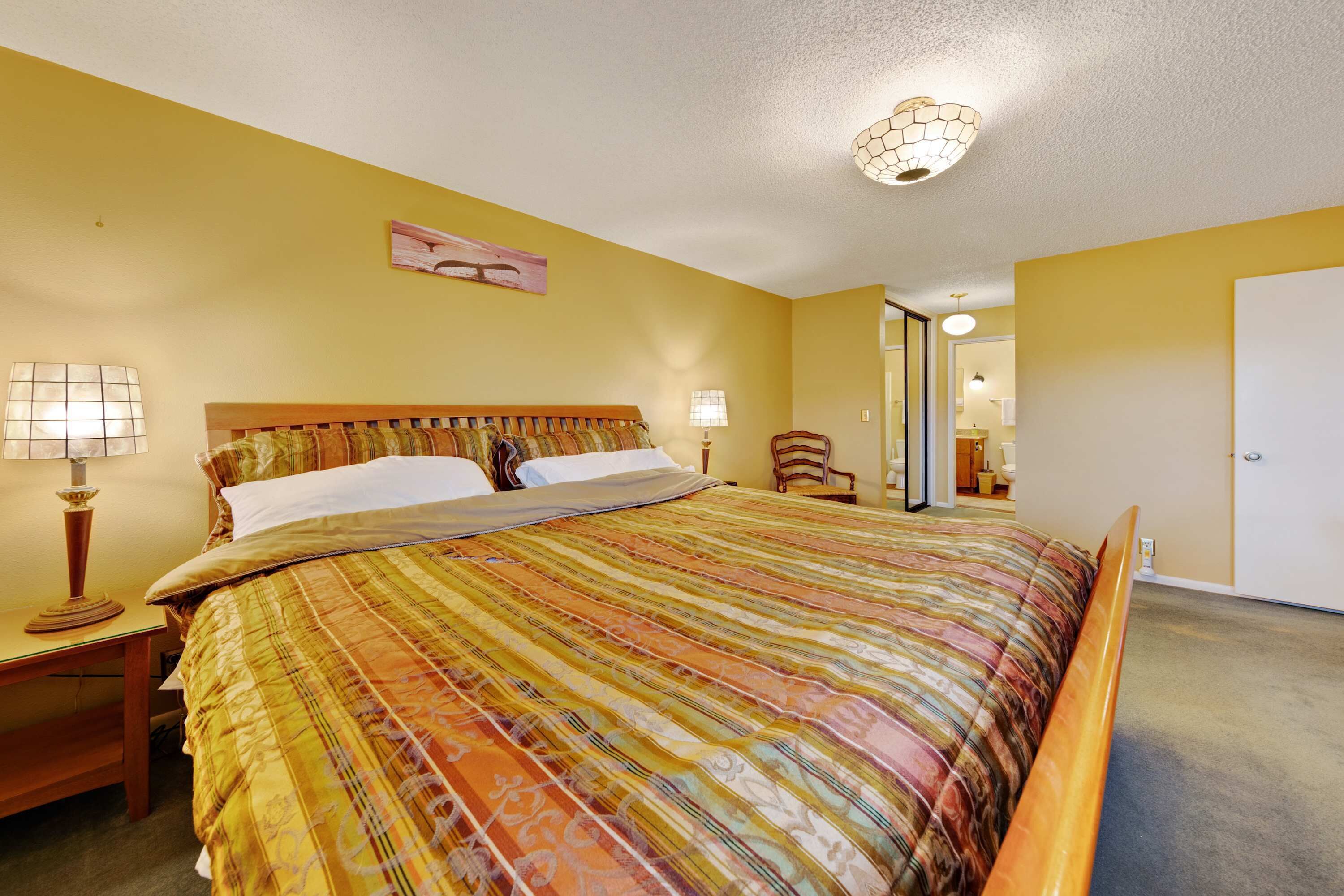 Ein Schlafzimmer mit einem großen Bett, zwei Nachttischlampen, einem hölzernen Kopfteil und einer Tür zu einem anderen Raum.