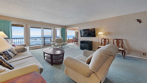 A spacious living room with a large window overlooking the sea, a flat-screen TV mounted on the wall, and a cozy seating area with a round coffee table. - Lovely oceanfront condo with sweeping water views and balcony (Gleneden Beach)