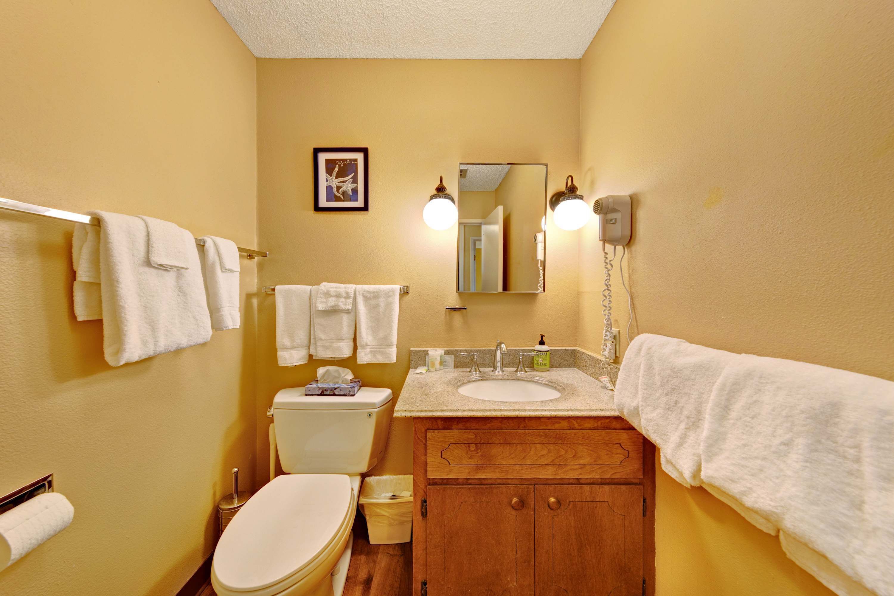 A bathroom with a toilet, sink, and wooden vanity. There are towels hanging on the wall and a framed picture above the mirror.