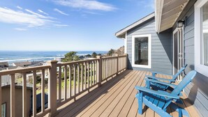 Una terraza de madera con vista al océano, una silla Adirondack azul y una casa con puerta blanca.