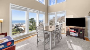 Un comedor con una mesa y sillas, un televisor sobre un mueble bajo, y ventanas amplias que ofrecen vista al océano y edificios circundantes.