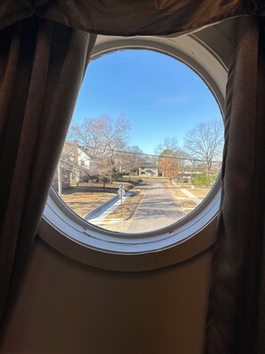Una ventana redonda con cortinas, a través de la cual se ve una calle con casas y árboles.