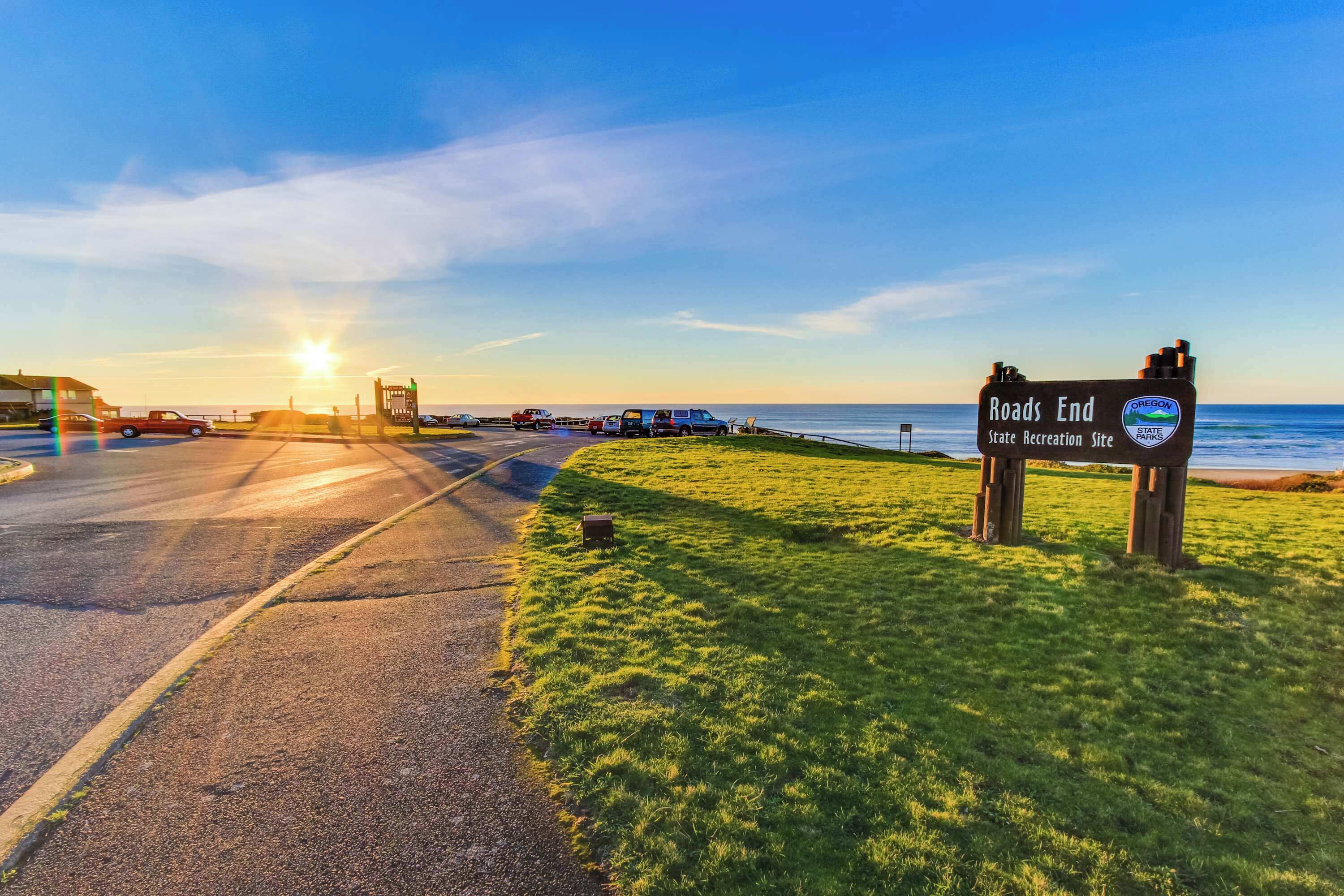 A road sign indicating 'Roads End' with a view of the ocean and parked vehicles.