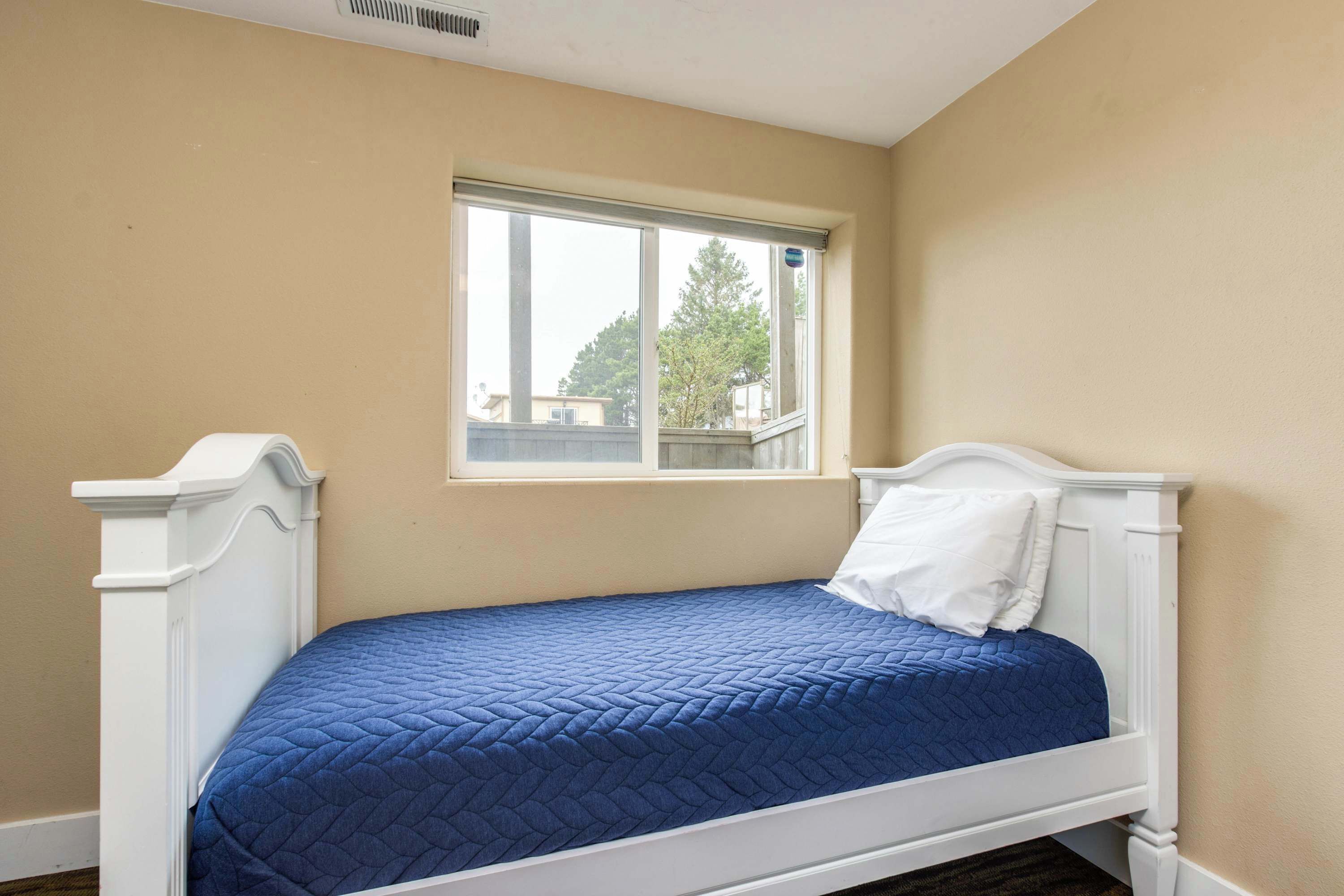 A single bed with a blue quilt in a room with beige walls and a window.