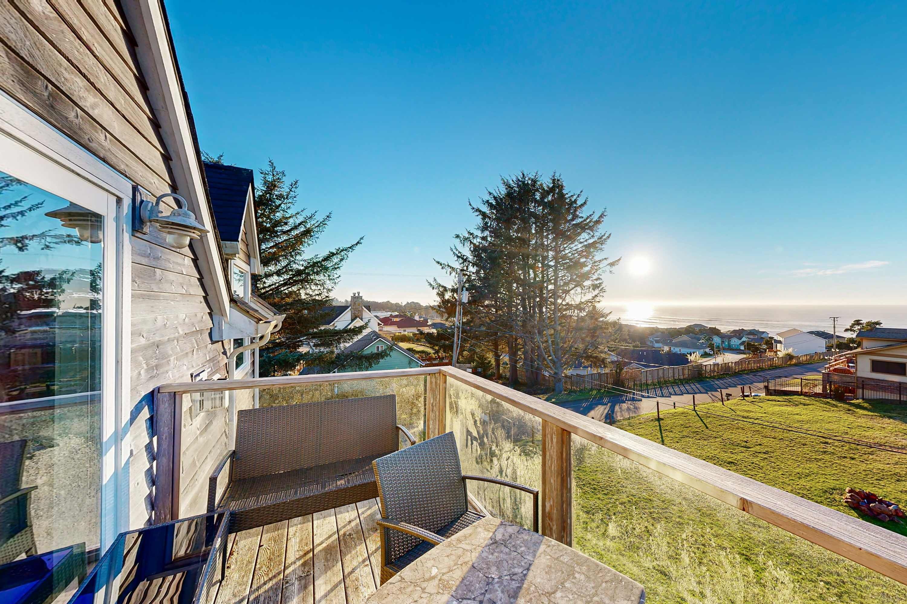 A wooden deck with a glass balustrade, outdoor seating, and a view of a grassy yard and distant houses.