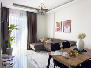 A modern living room with a sectional sofa, a dining table with chairs, a potted plant, and a vase of flowers. - Apartment 1+1 Qoople Saga 26 by Cleopatra Beach (Alanya)