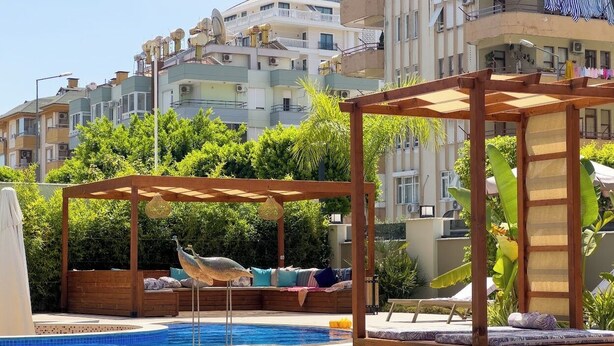 En swimmingpool med träpergola och avkopplande sittområden, omgiven av frodig grönska och bostadshus i bakgrunden.
