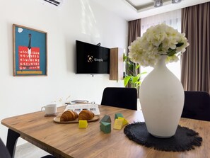 A dining table with croissants, a white vase with flowers, and a flat-screen TV on the wall. - Apartment 1+1 Qoople Saga 26 by Cleopatra Beach (Alanya)
