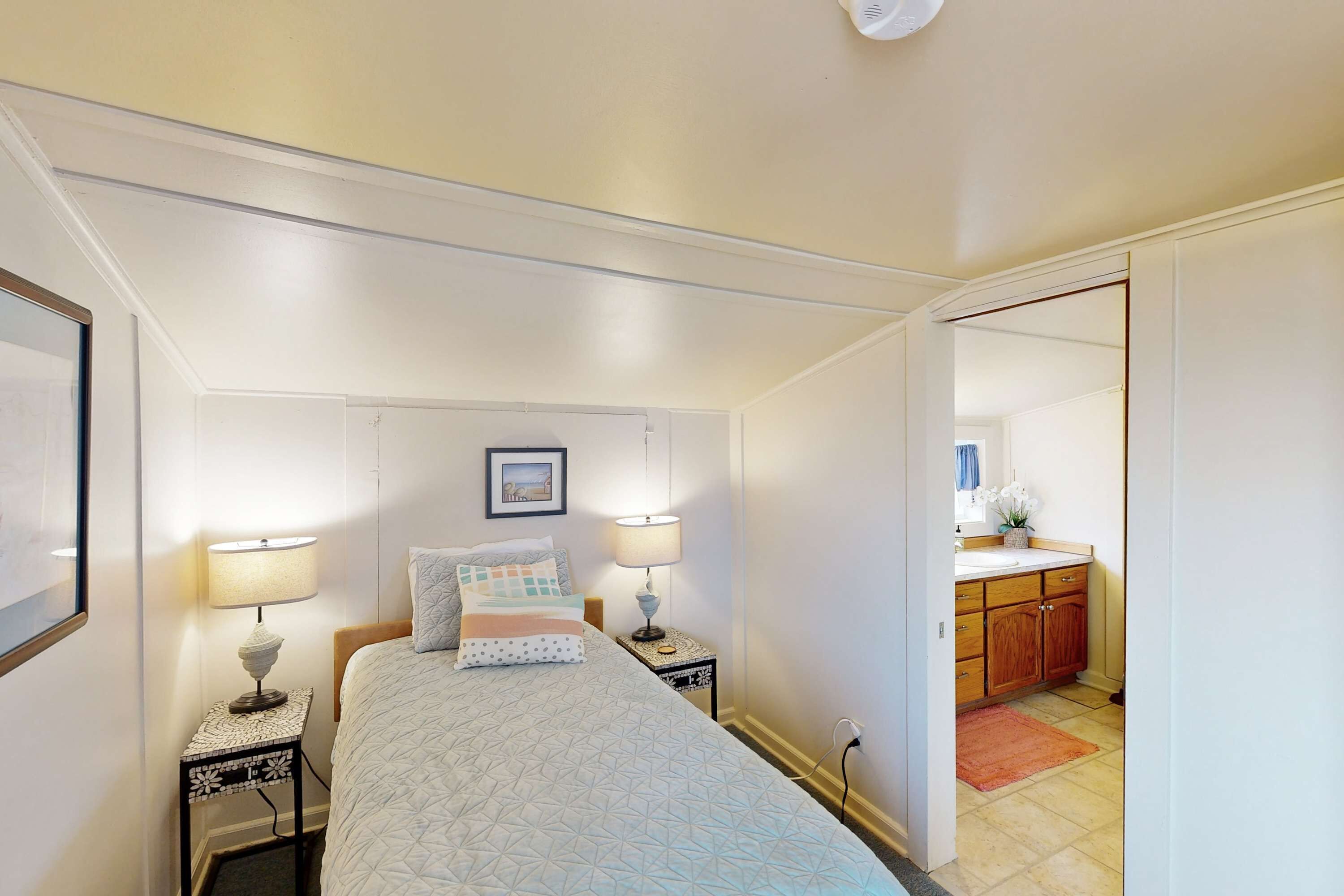 Un dormitorio con una cama, dos mesitas de noche con lámparas, una mesita auxiliar y una fotografía enmarcada en la pared, además de vistas al baño contiguo.