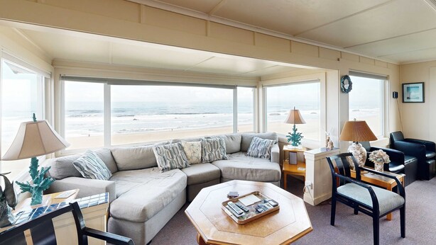 Een ruime woonkamer met een groot raam met uitzicht op het strand, een hoekbank, een salontafel met afstandsbedieningen en een bureau met een lamp.