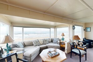 Amplia sala de estar con un ventanal con vista a la playa, un sofá seccional, una mesa de centro con controles remotos y un escritorio con una lámpara.
