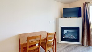 Una habitación con una mesa de comedor de madera y sillas, un televisor de pantalla plana sobre un mueble junto a la chimenea, y una cortina en la ventana.