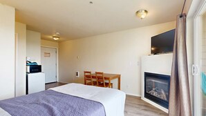 Habitación de hotel con cama, chimenea, televisor sobre la chimenea, mesa de comedor con sillas y área de cocina visible a través de una puerta abierta.