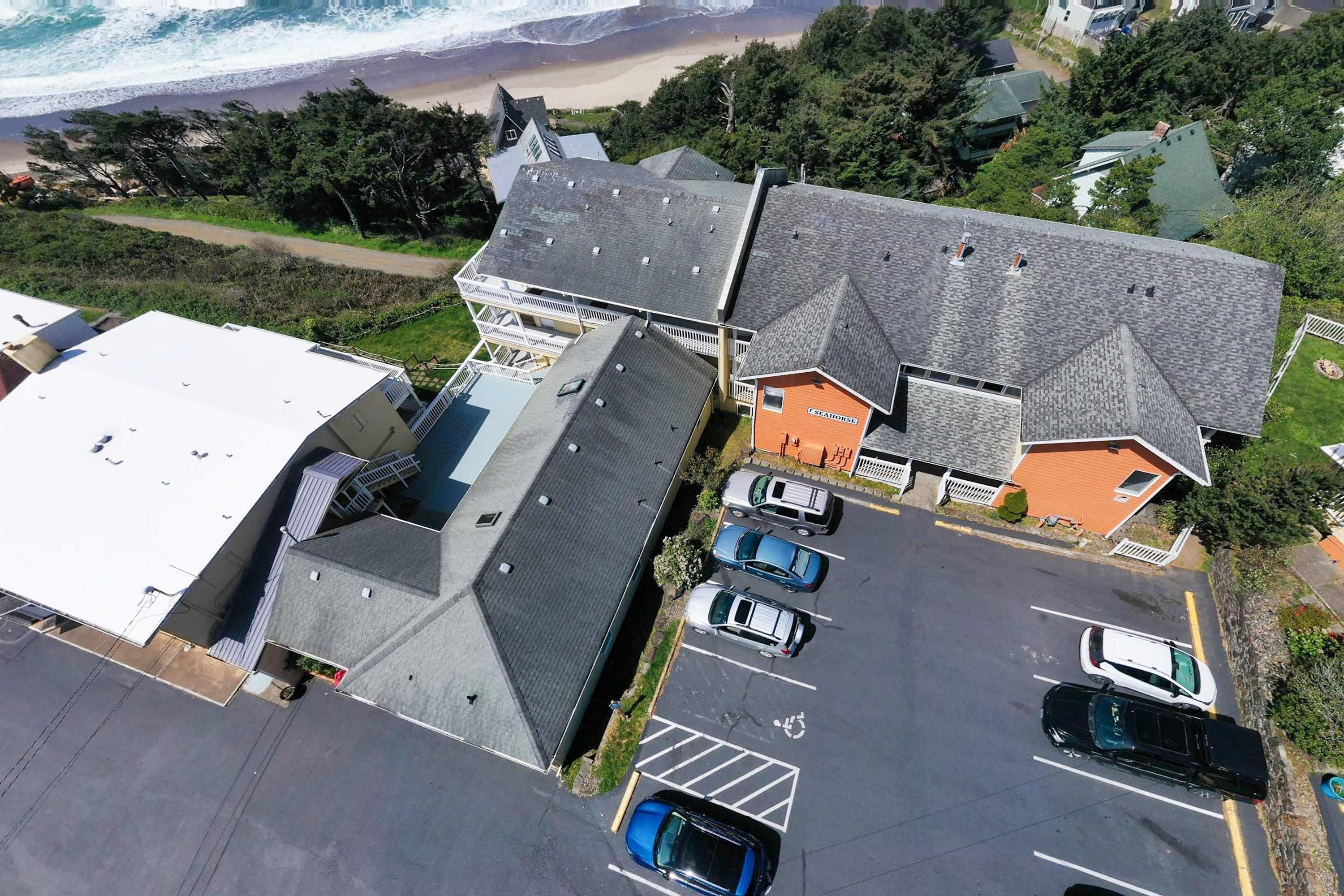 Une vue aérienne d’une propriété située sur la côte, avec plusieurs bâtiments, un parking où sont garées plusieurs voitures, et une plage en arrière-plan.
