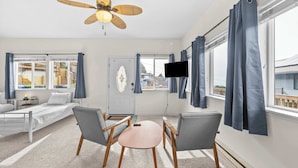 Une chambre d’hôtel comprenant un lit, deux fauteuils, une petite table, un ventilateur de plafond, un téléviseur fixé au mur et de grandes fenêtres avec des rideaux.