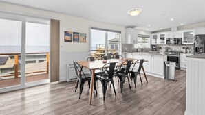Une cuisine moderne avec des meubles blancs, une table à manger en bois et des chaises noires. Une grande fenêtre offre une vue sur l’océan.