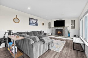 A modern living room with a grey sectional sofa, a fireplace, a TV mounted above it, and a large window providing natural light. - Gorgeous Retreat with Separate Cottage Private Hot Tub and Close Beach Access (Lincoln City)