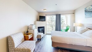A hotel room with a bed, a sofa, a TV, a fireplace, and a balcony with a chair. - Lower Level Ocean View Studio Close to the Beach Dogs Welcome (Lincoln City)